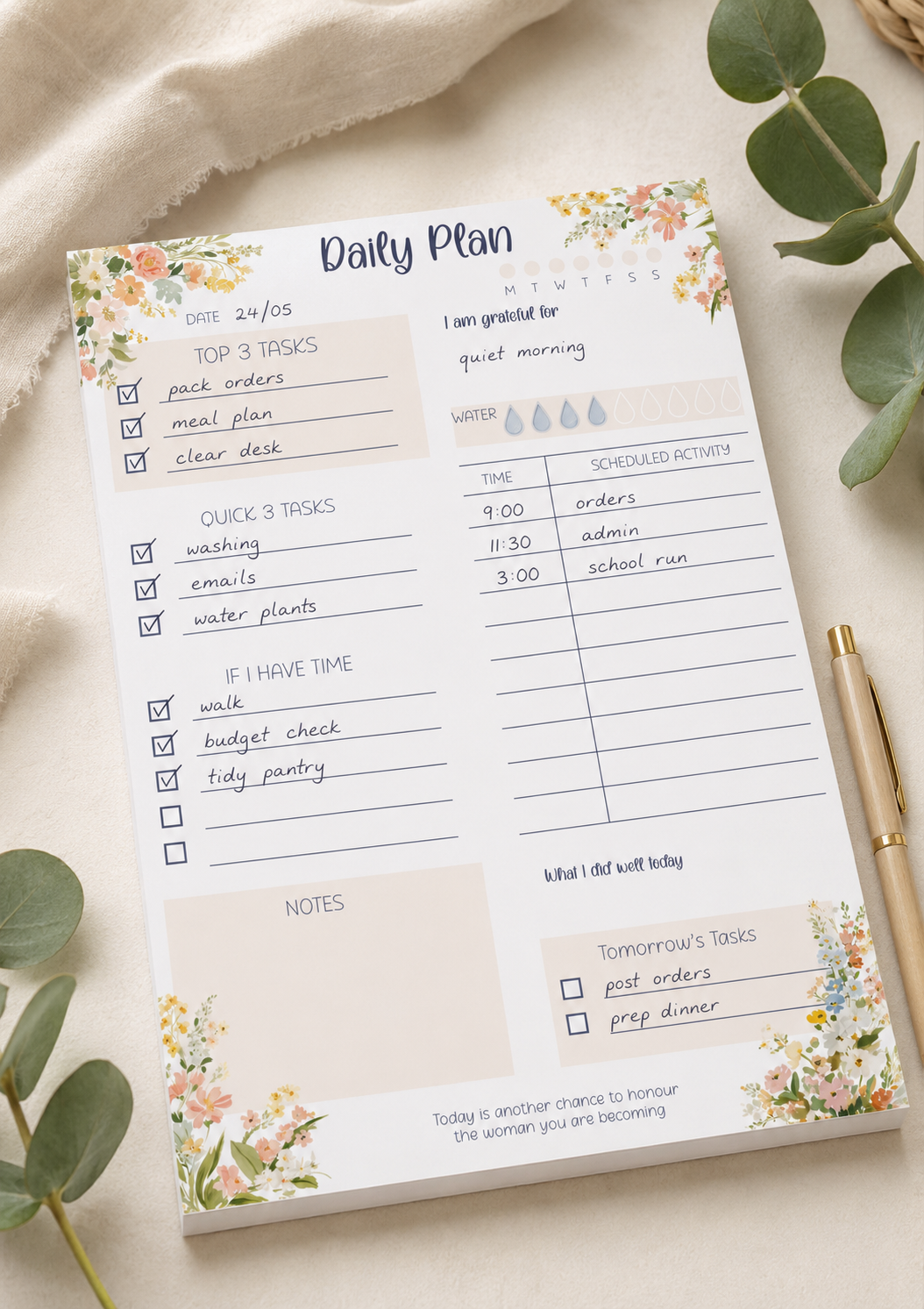 A floral 'Daily Plan' notepad sheet laid flat on a light fabric surface, surrounded by green eucalyptus leaves and a gold pen. The planner page includes sections for tasks, water intake, notes, and tomorrow's tables with pastel watercolor flowers at the corners.