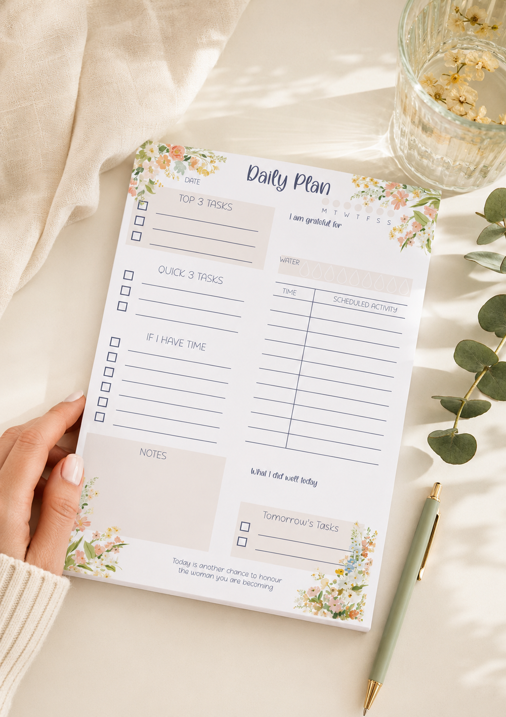 A hand holds a floral 'Daily Plan' notepad on a warm beige fabric surface, styled with a green pen, a glass of water, and eucalyptus leaves.