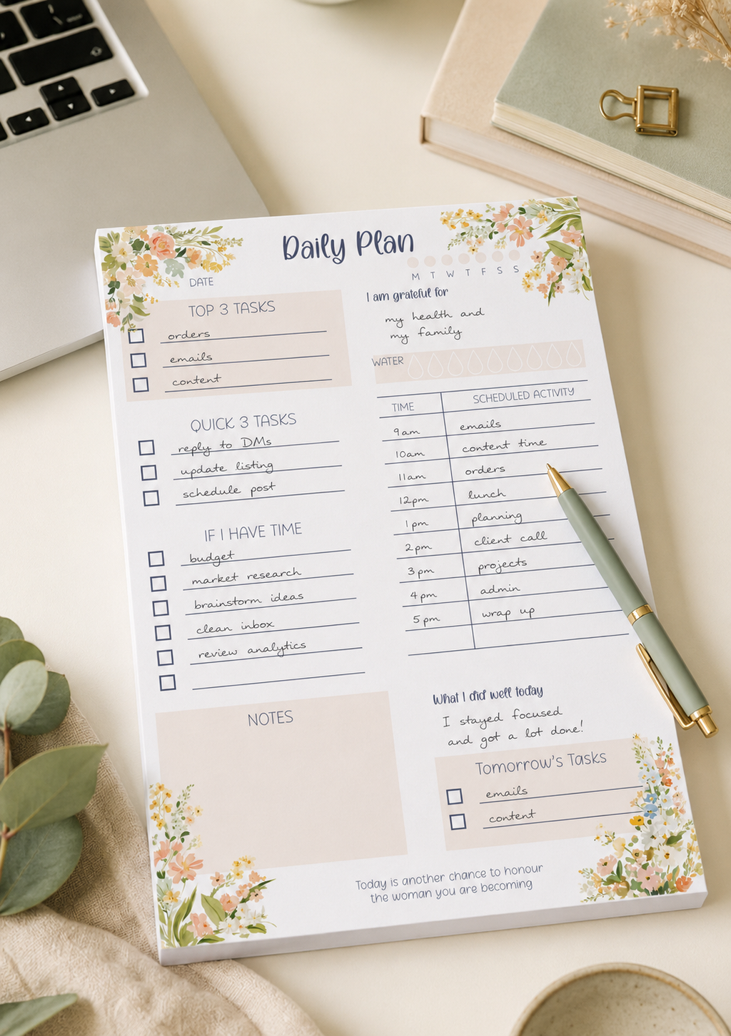 A floral 'Daily Plan' notepad sheet laid on a light beige surface, with a gold pen beside it. The scene includes part of a laptop, a small plant, and a decorative object in the background, making this a styled flat-lay rather than a plain studio product shot.