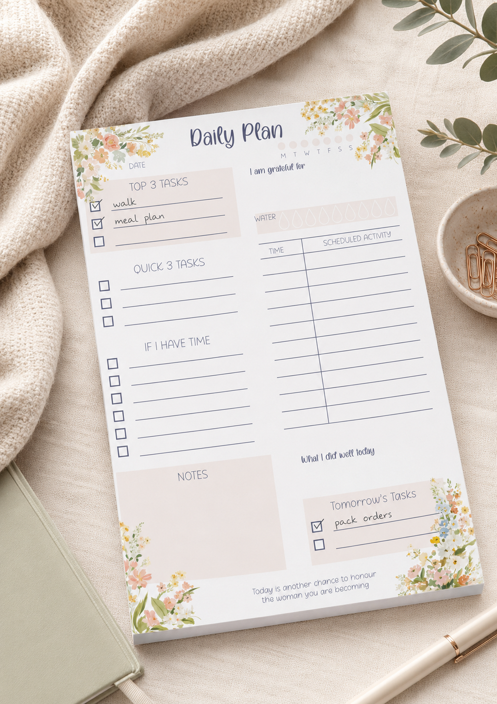 A floral 'Daily Plan' notepad sheet laid on a light beige fabric surface with a knit blanket, a small dish of gold coins, and a pen nearby. The notepad has watercolor-style flowers and is labeled 'Daily Plan' at the top.