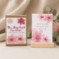 A boxed set of affirmation cards labeled 'The Abundant Creative Affirmation Cards' by Wanderful Gift Co, shown beside a single card on a small wooden stand. The packaging and card design feature pink watercolor flowers on a white background, with a soft beige fabric backdrop and eucalyptus leaves.