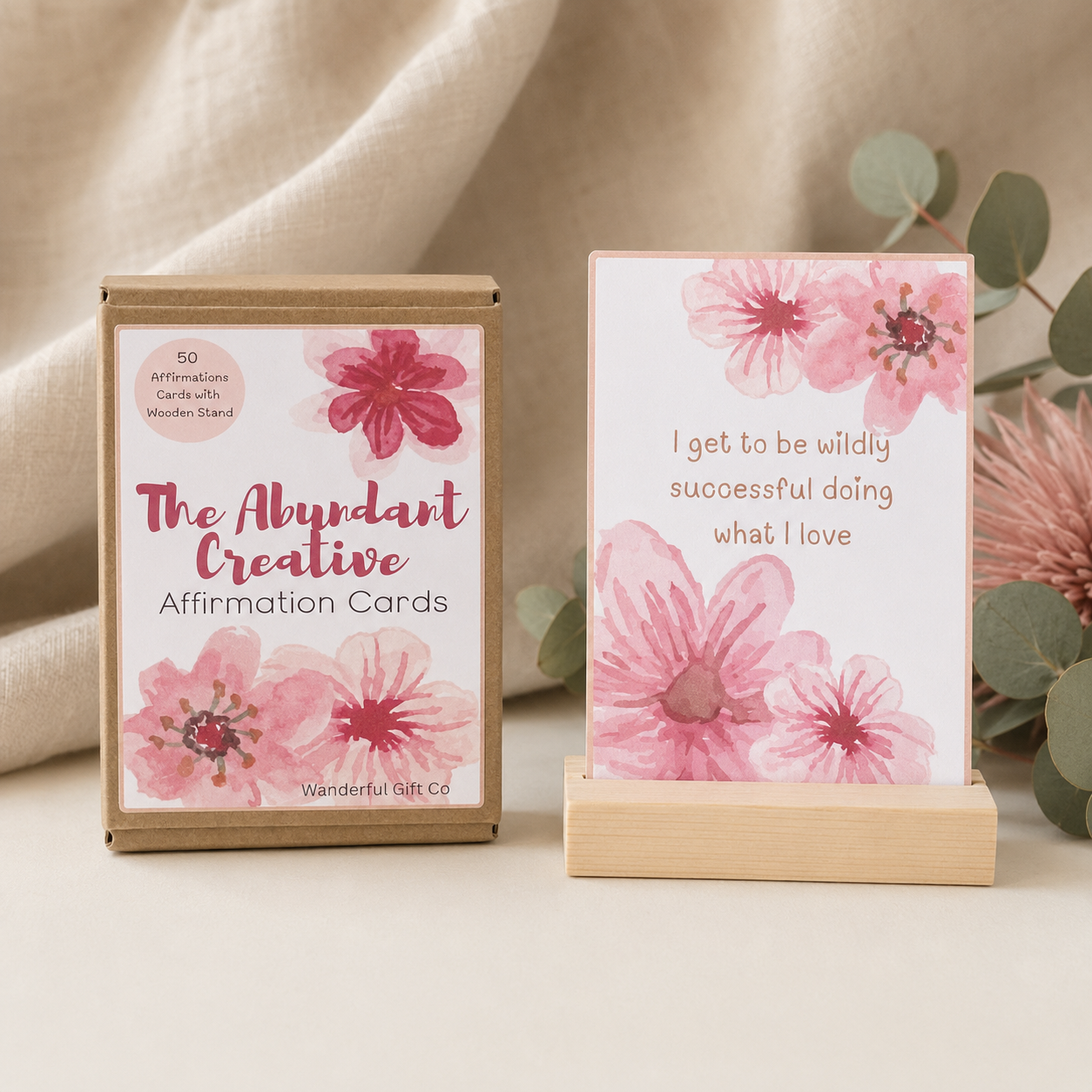 A boxed set of affirmation cards labeled 'The Abundant Creative Affirmation Cards' by Wanderful Gift Co, shown beside a single card on a small wooden stand. The packaging and card design feature pink watercolor flowers on a white background, with a soft beige fabric backdrop and eucalyptus leaves.