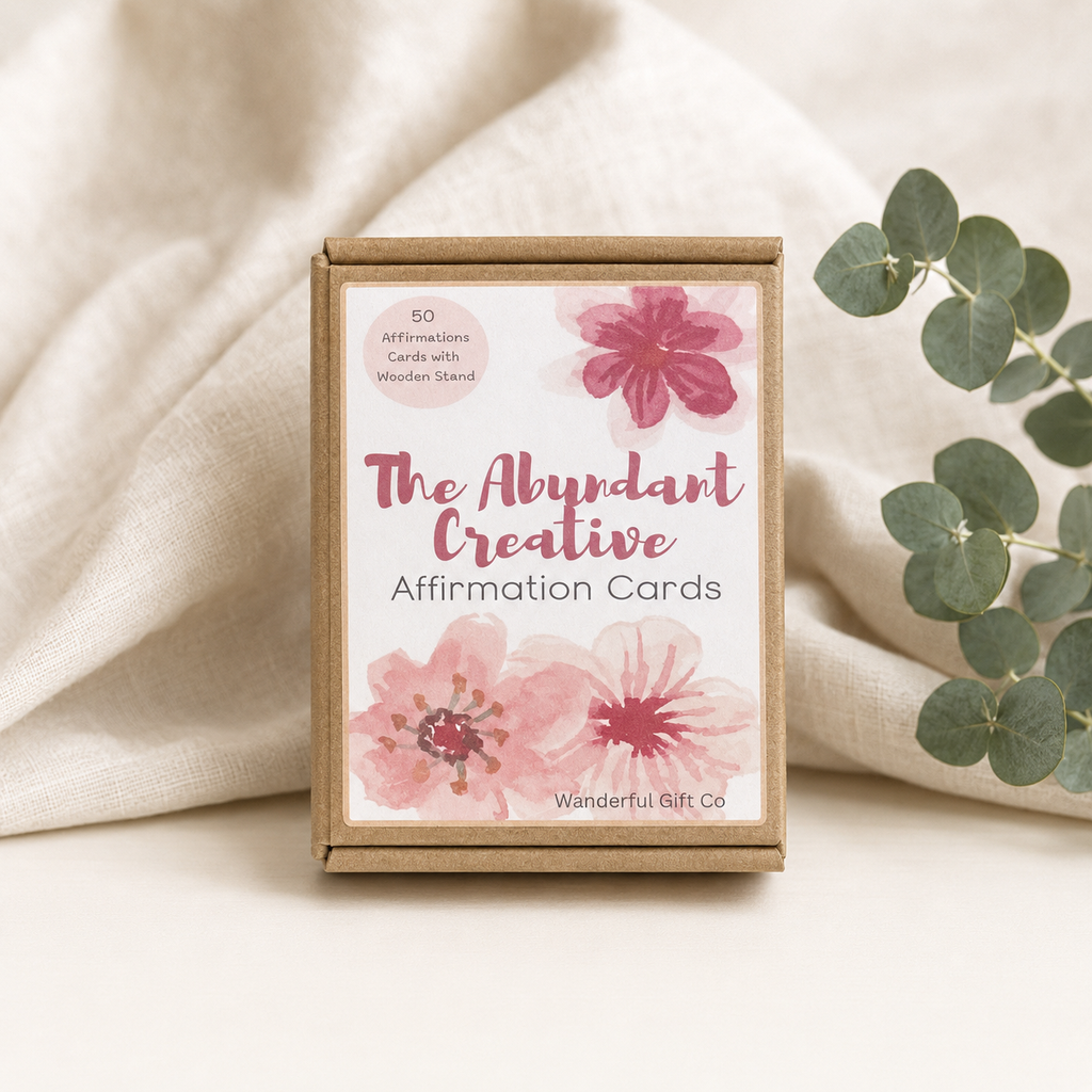 A kraft paper box of affirmation cards labeled 'The Abundant Creative Affirmation Cards' by Wanderful Gift Co, styled on a cream fabric surface with eucalyptus leaves.