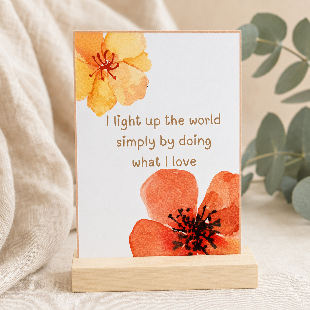 A vertical art print with a wooden stand, featuring a white background with watercolor orange and yellow flowers and the quote 'I light up the world simply by doing what I love' in brown text. The print is styled on a neutral fabric surface with soft eucalyptus leaves in the background.