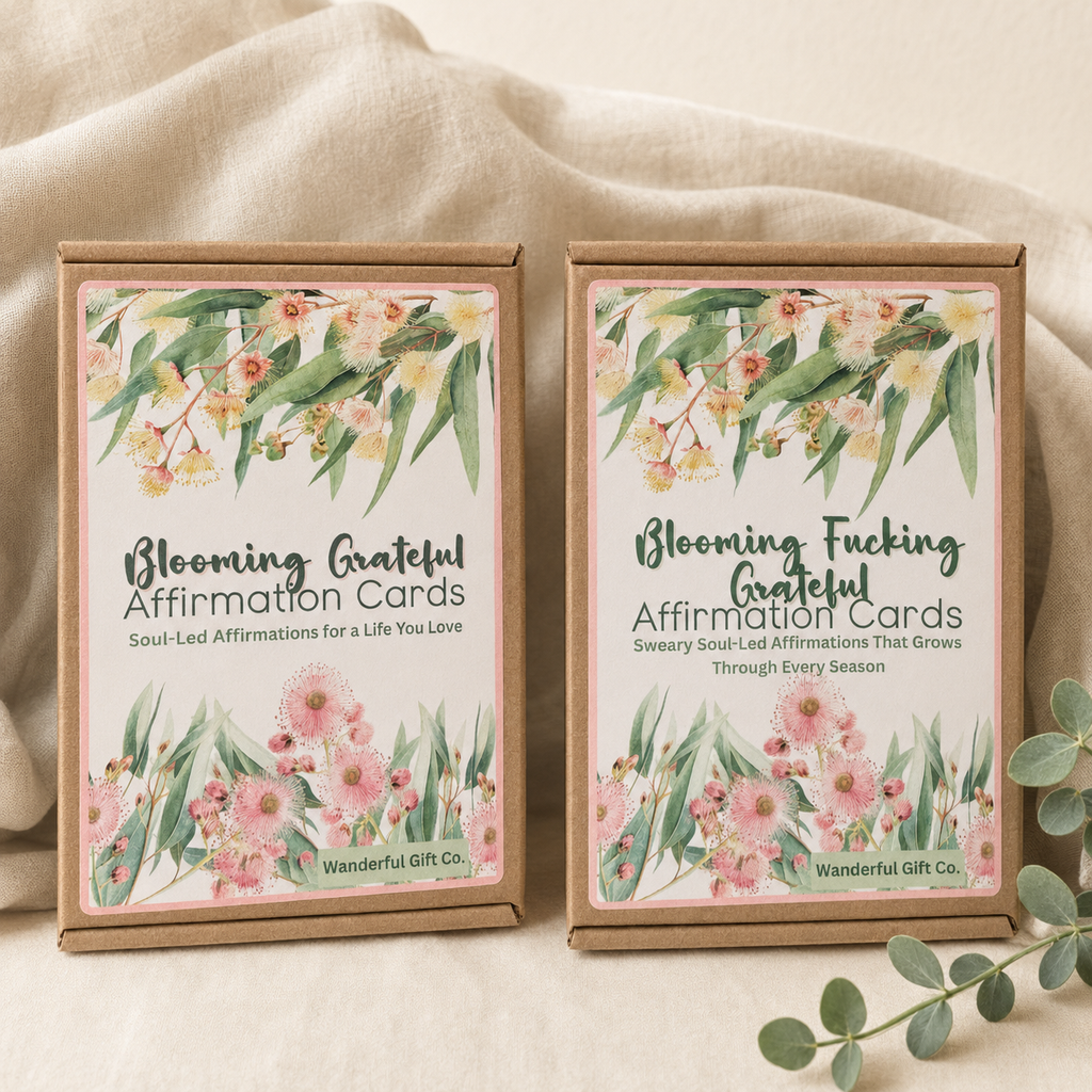Two boxed affirmation cards on a soft beige fabric background. The boxes are kraft paper with floral watercolor labels and readable text including 'Blooming Grateful Affirmation Cards' and 'Blooming Fucking Grateful Affirmation Cards,' with 'Wanderful Gift Co.