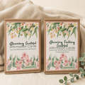 Two boxed affirmation cards on a soft beige fabric background. The boxes are kraft paper with floral watercolor labels and readable text including 'Blooming Grateful Affirmation Cards' and 'Blooming Fucking Grateful Affirmation Cards,' with 'Wanderful Gift Co.