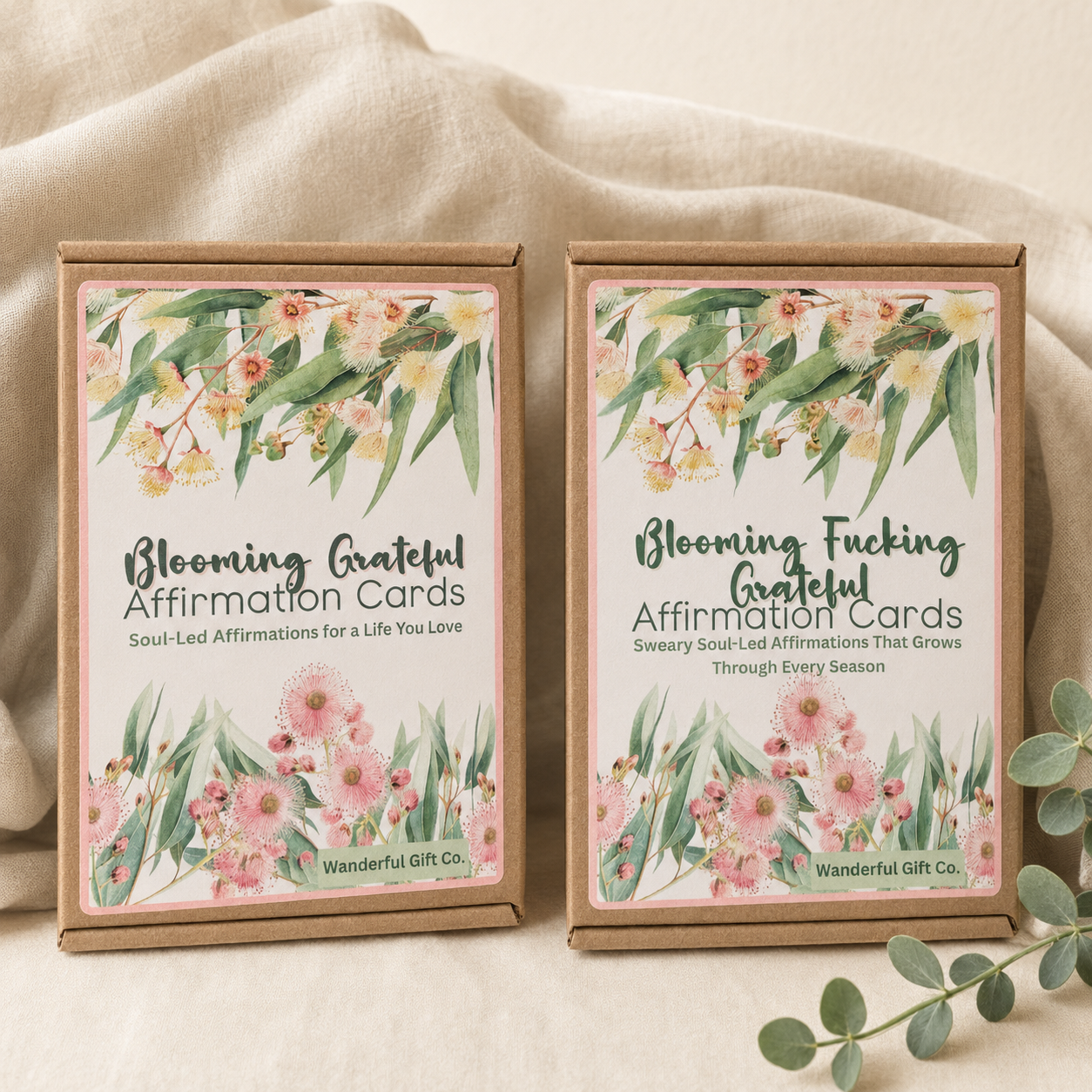 Two boxed affirmation cards on a soft beige fabric background. The boxes are kraft paper with floral watercolor labels and readable text including 'Blooming Grateful Affirmation Cards' and 'Blooming Fucking Grateful Affirmation Cards,' with 'Wanderful Gift Co.