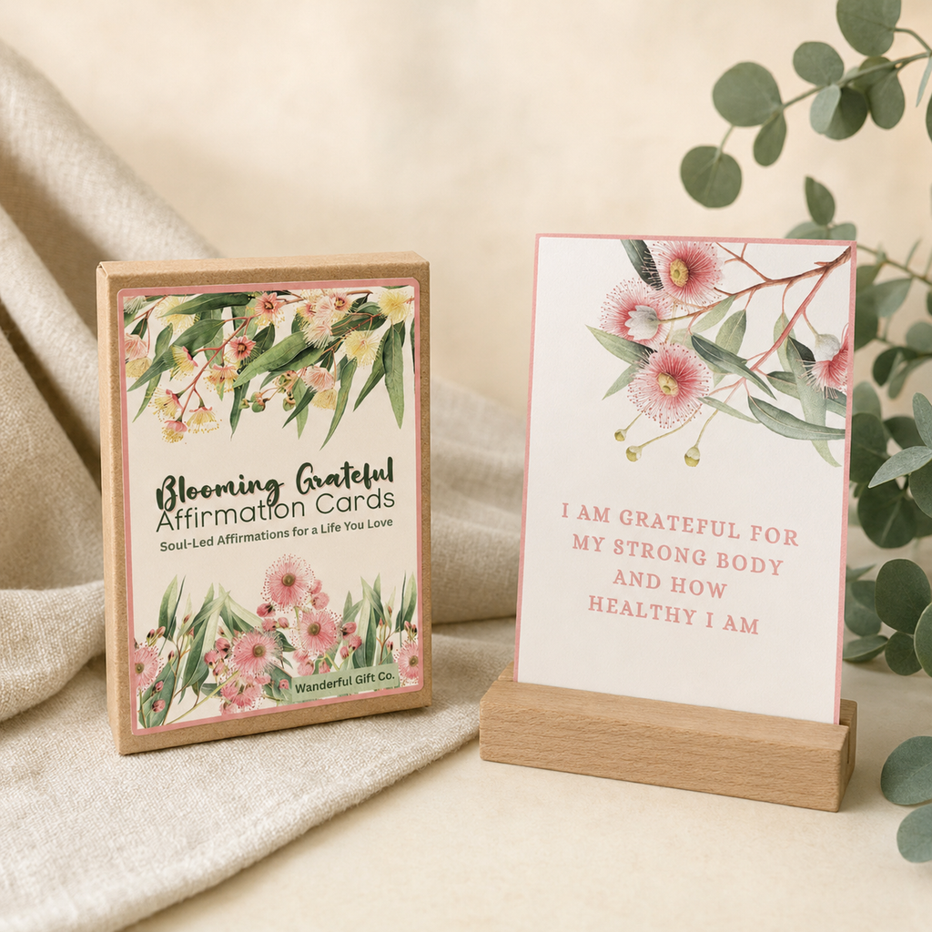 A boxed affirmation card set and a single card displayed on a wooden stand. The front of the box reads 'Blooming Grateful Affirmation Cards' with floral artwork, and the card says, 'I AM GRATEFUL FOR MY STRONG BODY AND HOW HEALTHY I AM.