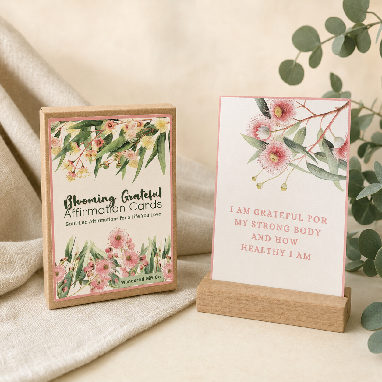 A boxed affirmation card set and a single card displayed on a wooden stand. The front of the box reads 'Blooming Grateful Affirmation Cards' with floral artwork, and the card says, 'I AM GRATEFUL FOR MY STRONG BODY AND HOW HEALTHY I AM.