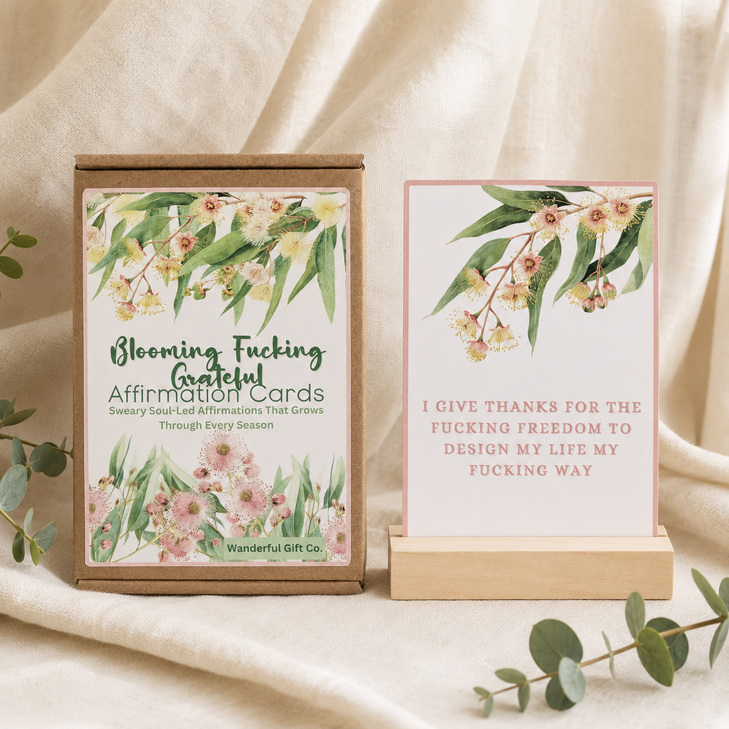 A boxed affirmation card set and a single standing card displayed on a draped cream fabric background with eucalyptus leaves. The box reads 'Blooming Fucking Grateful Affirmation Cards' and shows floral artwork; the card says, 'I give thanks for the fucking freedom to design my life my fucking way.