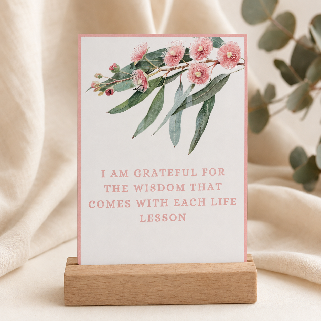 A vertical printed card or sign with a watercolor floral branch at the top and the text 'I AM GRATEFUL FOR THE WISDOM THAT COMES WITH EACH LIFE LESSON' in pink lettering, displayed on a small wooden stand against a soft beige fabric backdrop with blurred greenery.