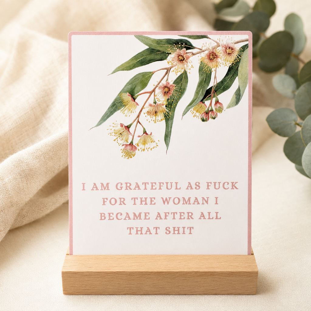 A vertical printed sign or card with a pink border, watercolor eucalyptus flowers and leaves at the top, and pink text reading 'I AM GRATEFUL AS FUCK FOR THE WOMAN I BECAME AFTER ALL THAT SHIT' displayed on a small wooden stand in a styled tabletop setting.