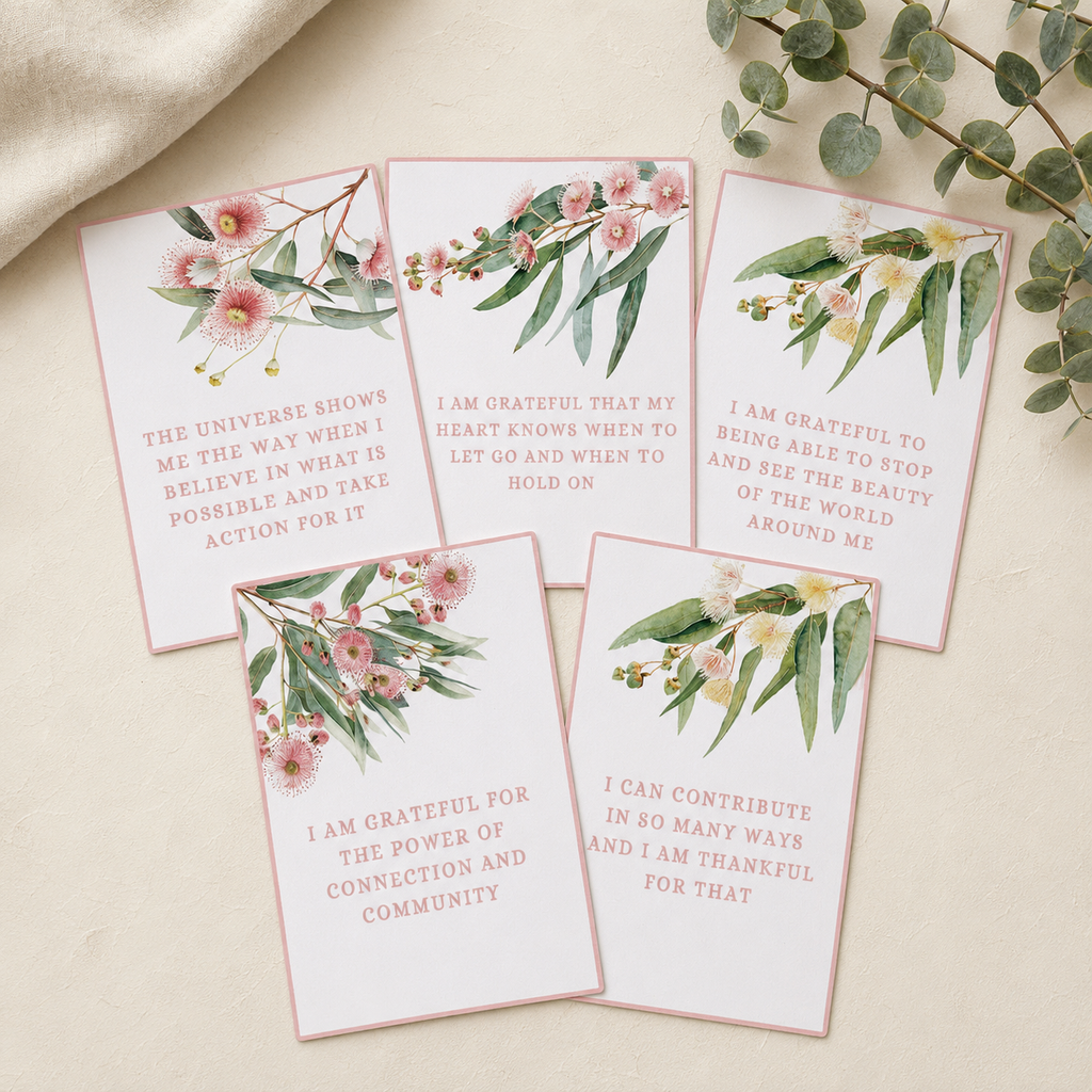Five rectangular printed cards with watercolor floral illustrations and inspirational quotes, arranged on a light beige surface with fabric and eucalyptus branches around them.