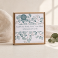 A square kraft paper box of affirmation cards sits on a light beige surface with soft fabric and eucalyptus leaves around it. The front label reads 'Fuel Your Body, Free Your Mind Affirmation Cards' with watercolor eucalyptus branches in muted blue-green tones.