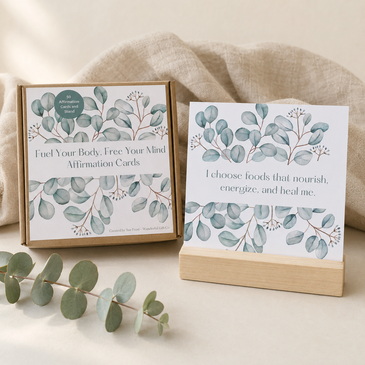  affirmation cards displayed in a styled setting with a kraft paper box, a wooden stand, and eucalyptus leaves on a neutral fabric background.