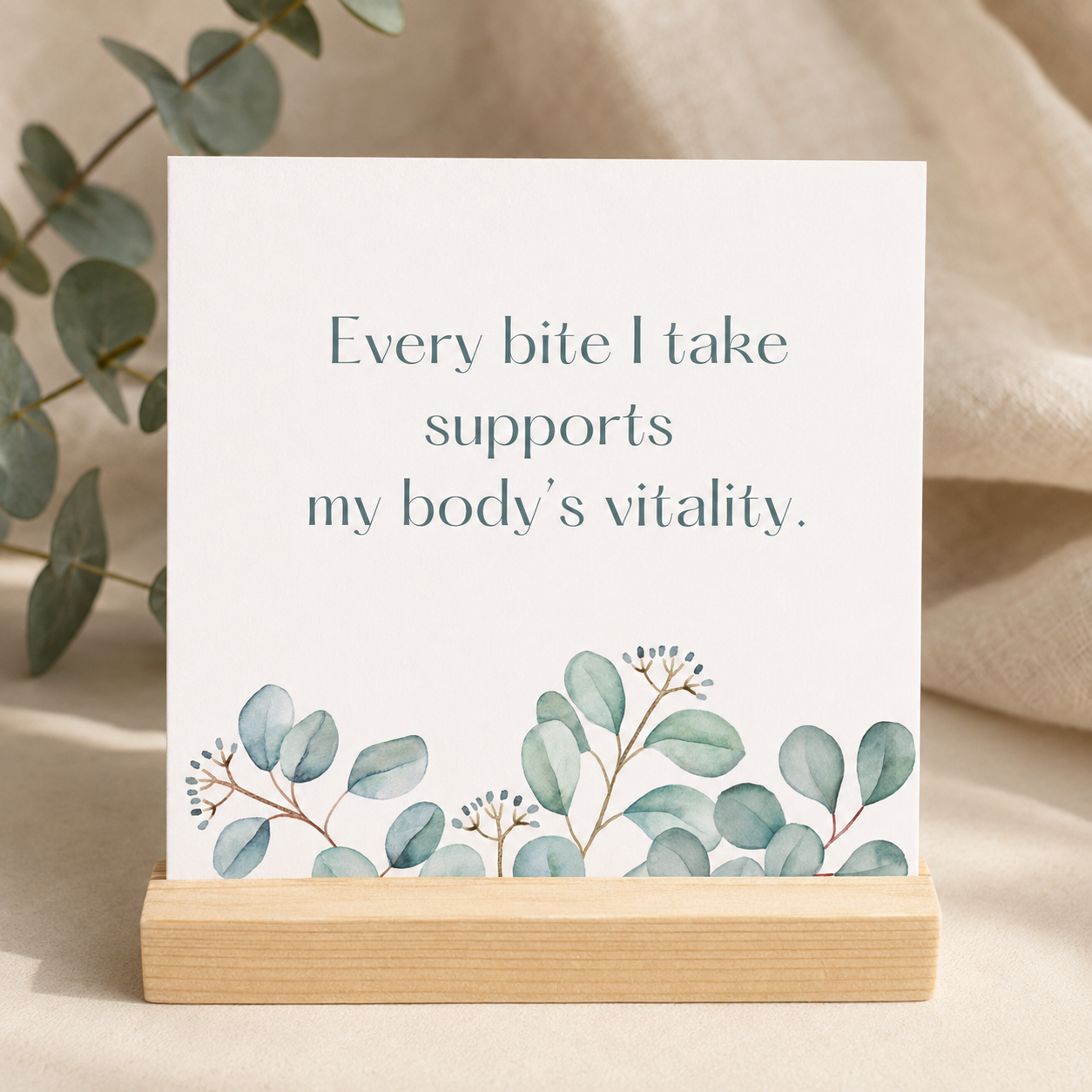 A square white sign or card on a light wood stand, printed with the phrase 'Every bite I take supports my body's vitality.' in muted blue-gray text and decorated with watercolor eucalyptus leaves along the bottom.