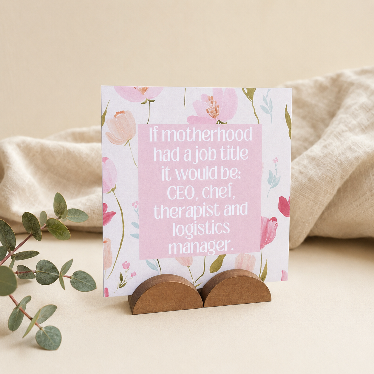 A floral greeting card with a pink text box reading, 'If motherhood had a job title it would be: CEO, chef, therapist and logistics manager.' displayed on a small wooden stand against a soft beige fabric backdrop with eucalyptus leaves in the foreground.
