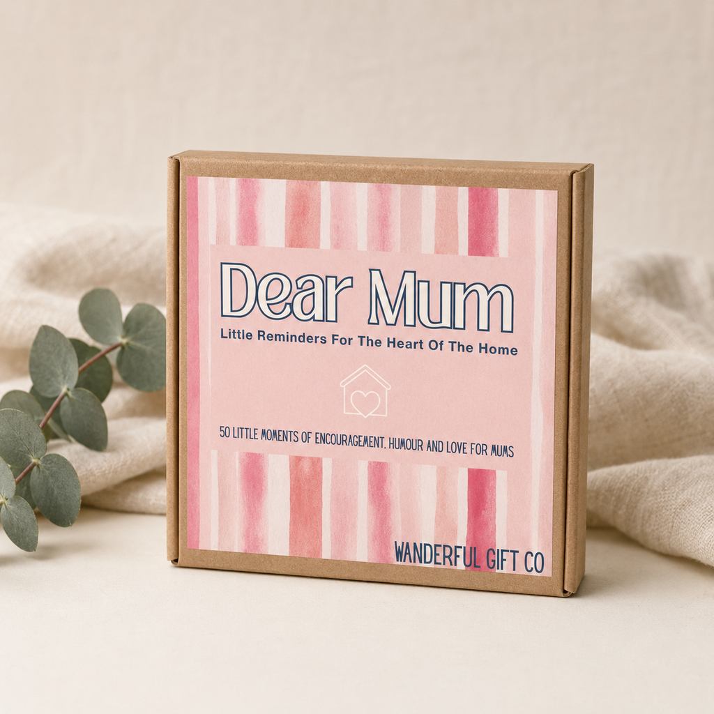 A square kraft gift box with a pink watercolor striped label reading 'Dear Mum' and 'WANDEREFUL GIFT CO', styled on a light beige surface with soft fabric and eucalyptus leaves around it.