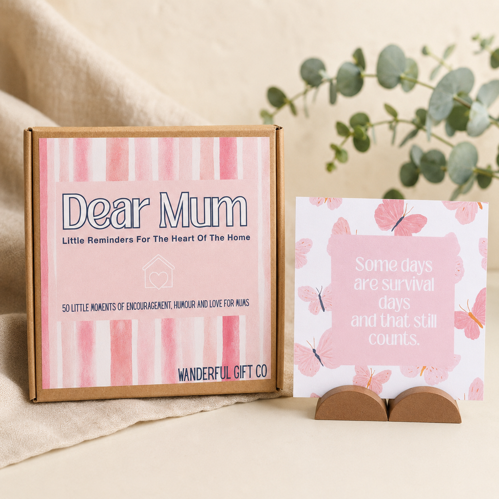 A gift set for 'Dear Mum' by Wonderful Gift Co, featuring a brown cardboard box with pink watercolor stripes and a matching pink butterfly-patterned card on a small wooden stand, styled on a beige fabric surface with eucalyptus branches in the background.