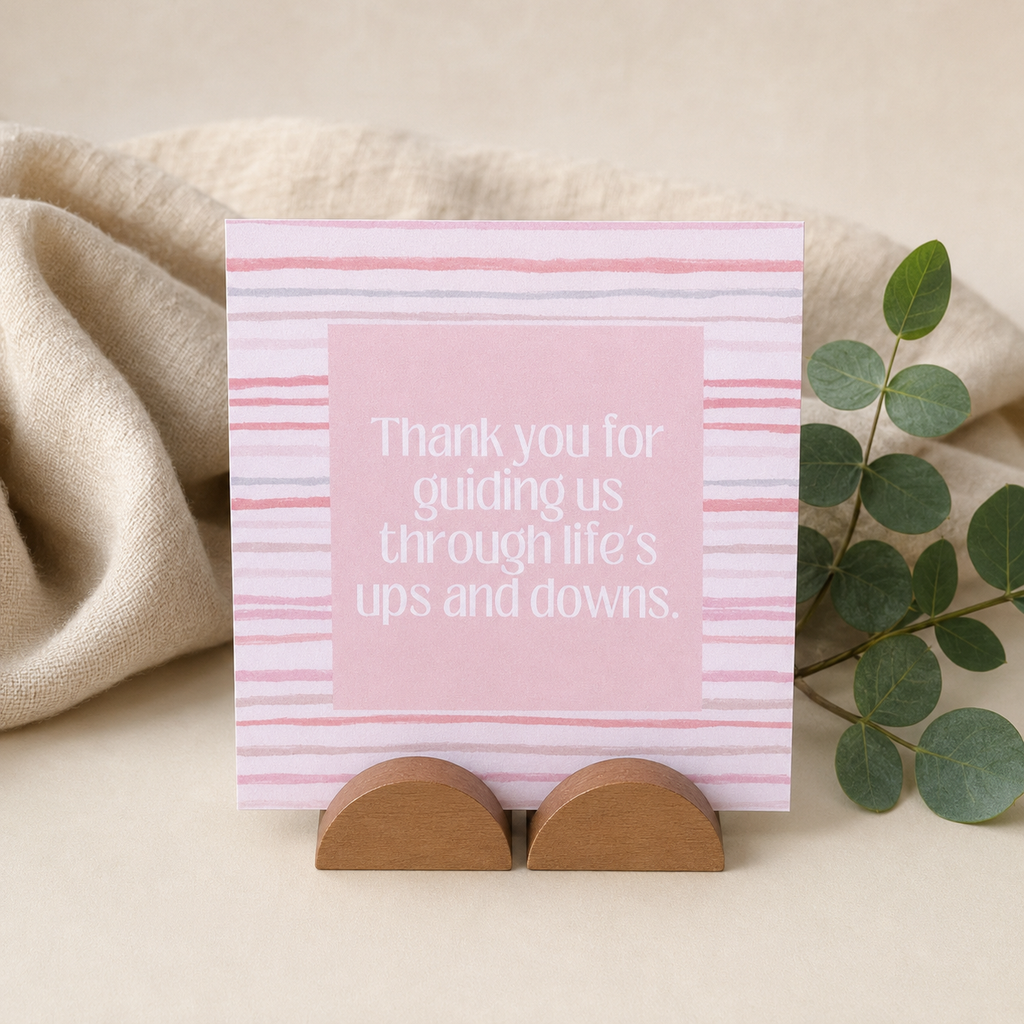 A square pink thank-you card with white text reading 'Thank you for guiding us through life's ups and downs.' stands on two small wooden display stands.