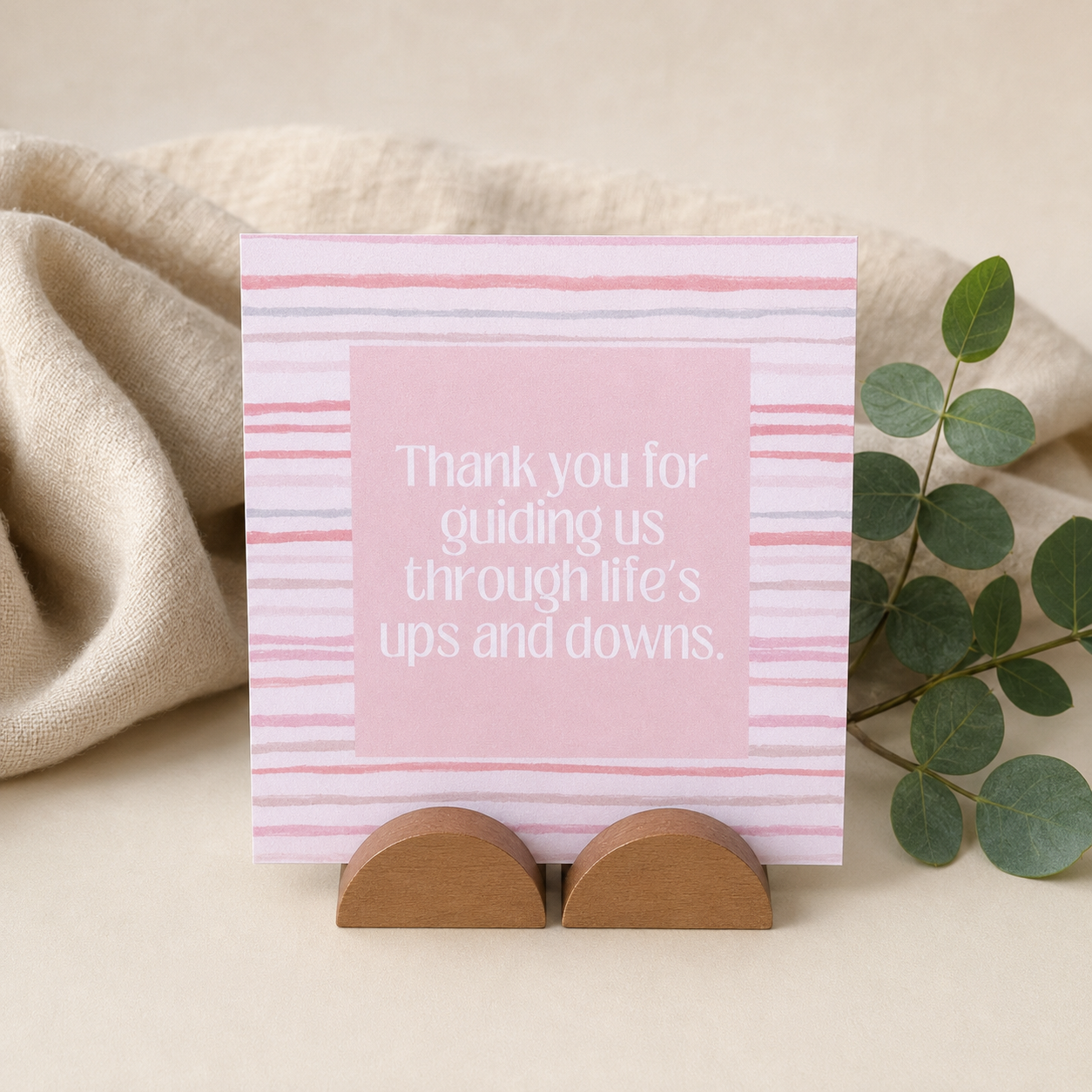 A square pink thank-you card with white text reading 'Thank you for guiding us through life's ups and downs.' stands on two small wooden display stands.