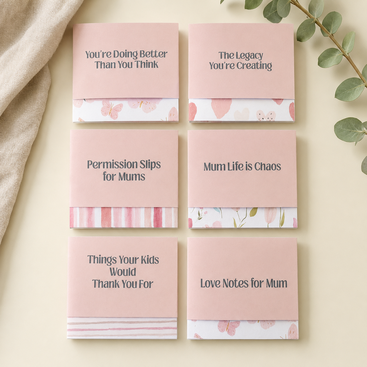 Six pink postcard-style cards with humorous Mother's Day messages are arranged in a 2x3 grid on a light cream background with a beige fabric corner and eucalyptus leaves. The cards feature different printed phrases and decorative pink floral and striped borders along the bottom.
