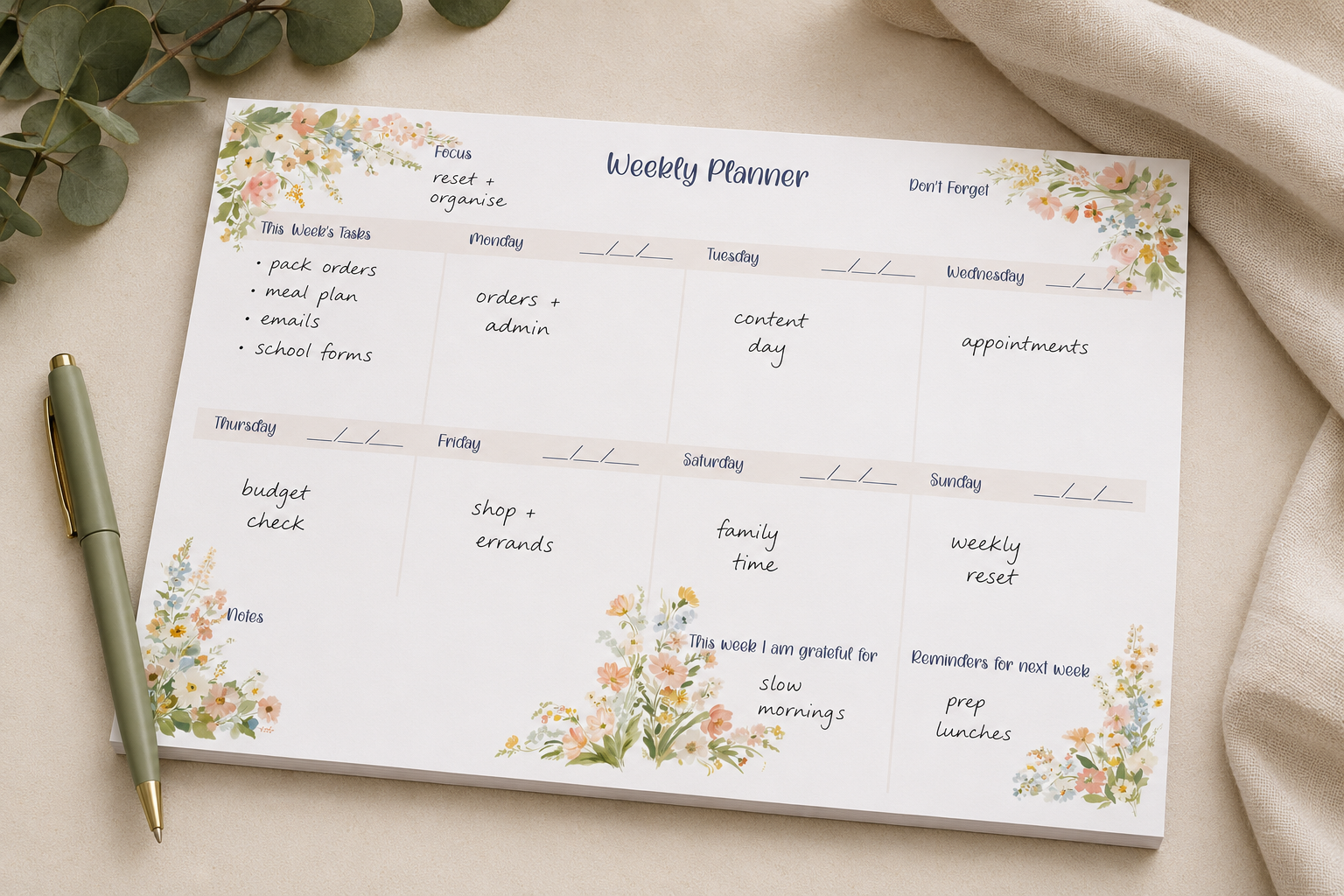 A floral 'Weekly Planner' notepad laid flat on a beige fabric surface, with a green pen beside it and eucalyptus leaves around the edges. The pad has watercolor-style flowers and leaves in soft pink, yellow, and green, with sections for each day of the week and handwritten notes.