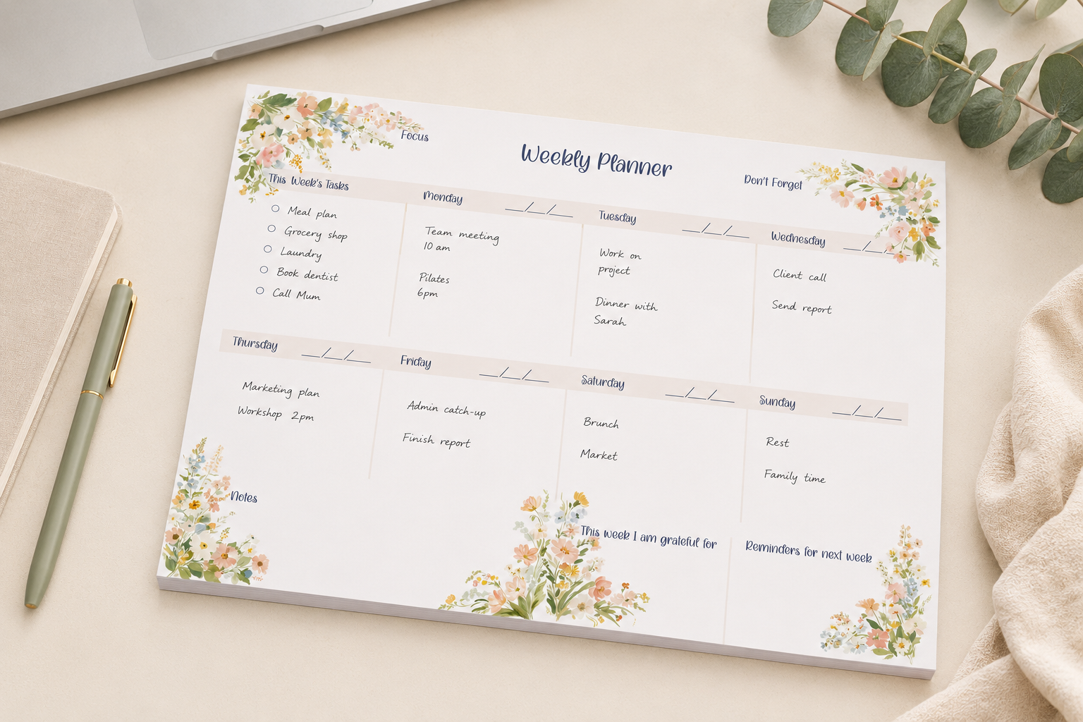 A weekly planner notepad with watercolor floral designs in the corners, styled on a light beige surface with a pen, notebook, and eucalyptus leaves.