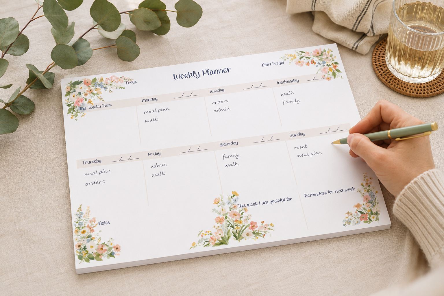 A weekly planner pad with floral watercolor designs at the corners, shown on a beige tabletop with a hand holding a green pen writing in one of the boxes. A glass of water, a folded napkin, and eucalyptus leaves are visible in the background.