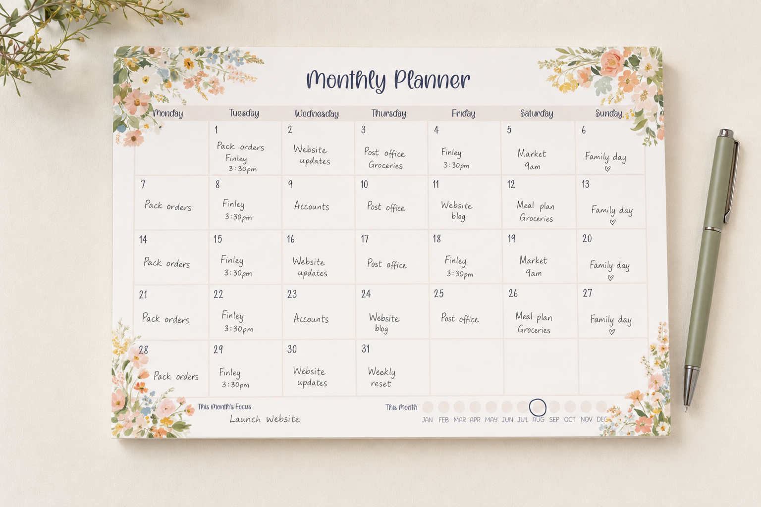 A floral monthly planner pad shown from above on a light beige surface, with a silver pen to the right and small greenery in the top corner. The planner has a white page with pastel peach and green watercolor flowers around the border and a printed calendar grid for the month.