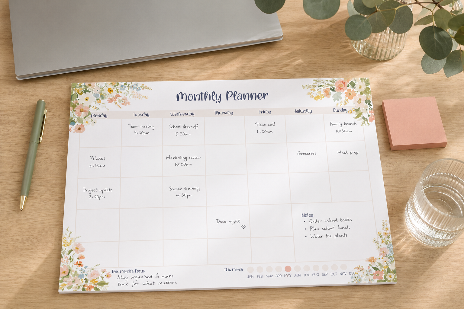 A floral monthly planner pad laid flat on a wooden desk, surrounded by a laptop, pen, sticky notes, glass, and greenery.