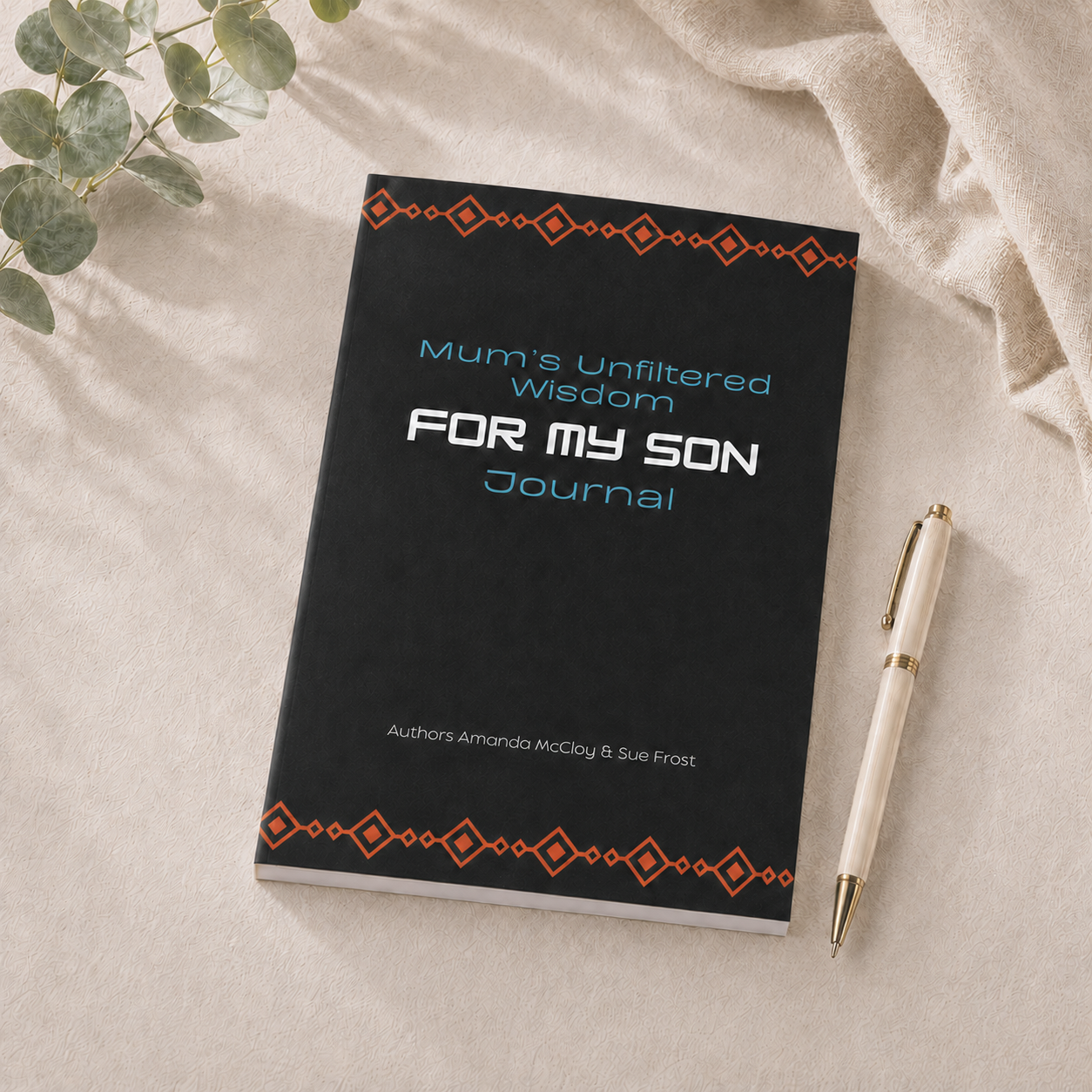 A black hardcover journal titled 'Mum's Unfiltered Wisdom FOR MY SON Journal' with orange geometric borders and light blue and white text, styled on a beige fabric surface with a cream pen and green leaves.