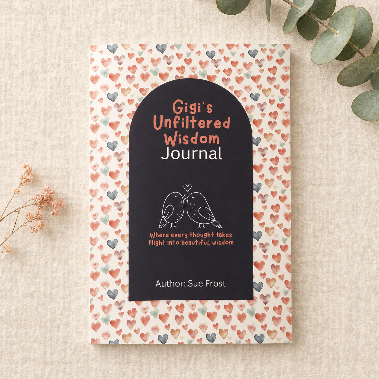 A journal cover shown flat on a light beige surface with decorative eucalyptus leaves and dried flowers. The cover has a white background covered in small pink, peach, and gray hearts, with a large black arch-shaped panel in the center containing the title 'Gigi's Unfiltered Wisdom Journal' and a simple bird illustration.