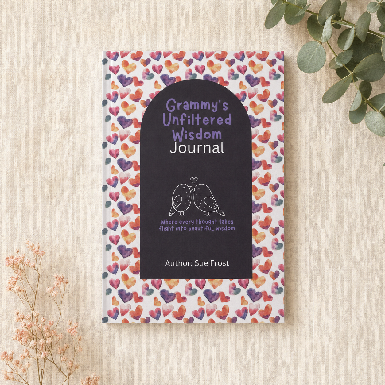 A hardcover journal titled 'Grammy's Unfiltered Wisdom Journal' by Sue Frost, featuring a black arched cover with white and purple text and bird illustrations, set against a pattern of colorful hearts. The book is styled on a textured beige fabric background with dried flowers and eucalyptus leaves.
