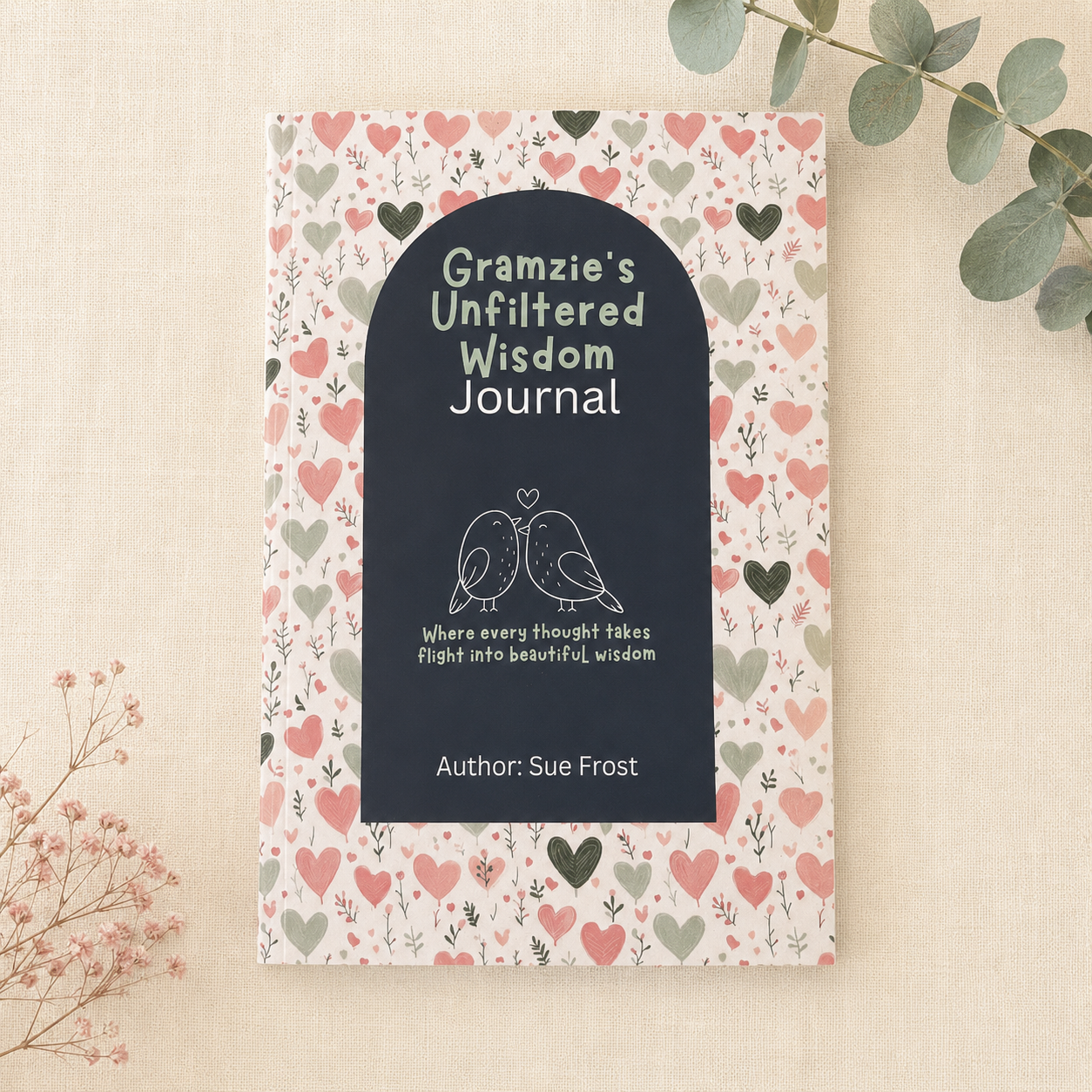 A journal titled 'Gramzie's Unfiltered Wisdom Journal' by Sue Frost, featuring a dark navy blue front cover with white text and a bird illustration, surrounded by a pink, red, and green heart patterned border. The journal is displayed flat on a textured beige fabric background with decorative eucalyptus and baby's breath flowers.