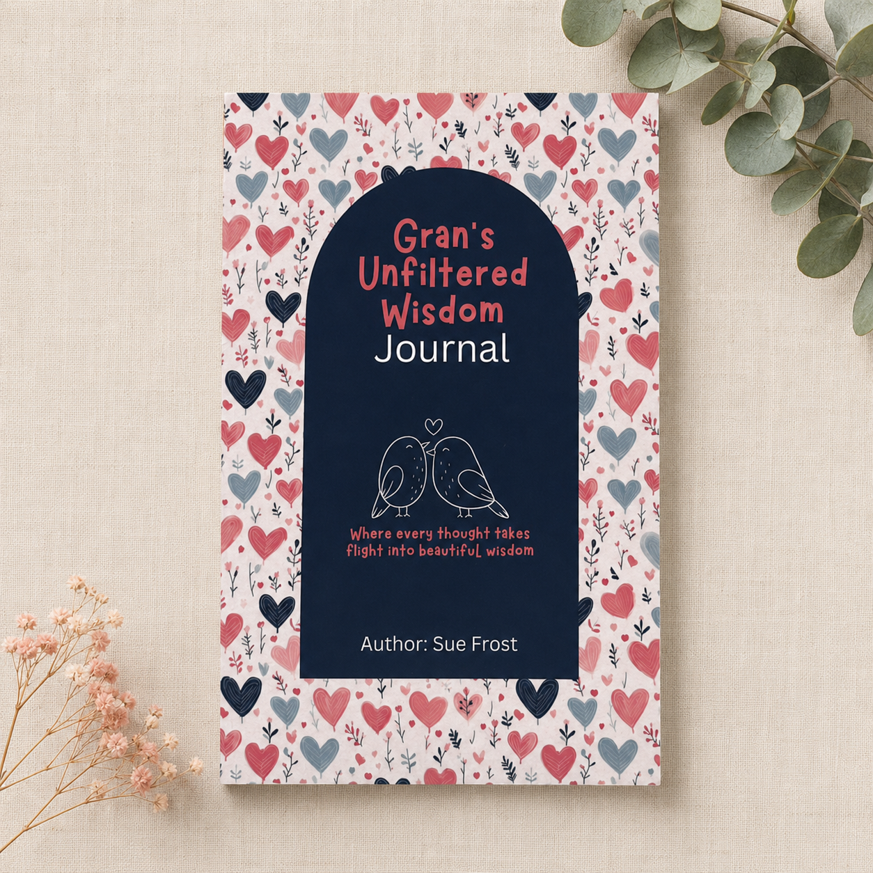A hardcover journal titled 'Gran's Unfiltered Wisdom Journal' by Sue Frost, featuring a navy blue arched center with white and coral text, surrounded by a pink, red, and blue heart pattern. The journal is displayed on a textured beige fabric background with decorative eucalyptus and dried flowers.