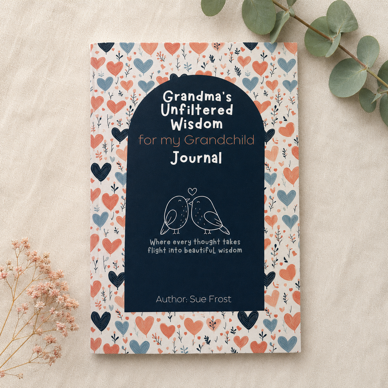 A journal cover laid flat on a light fabric surface, surrounded by decorative dried flowers and eucalyptus leaves. The cover has a cream background with a border of small hearts in coral, navy, and blue, plus a large dark navy central panel with white text reading 'Grandma's Unfiltered Wisdom for my Grandchild Journal' and 'Author: Sue Frost.