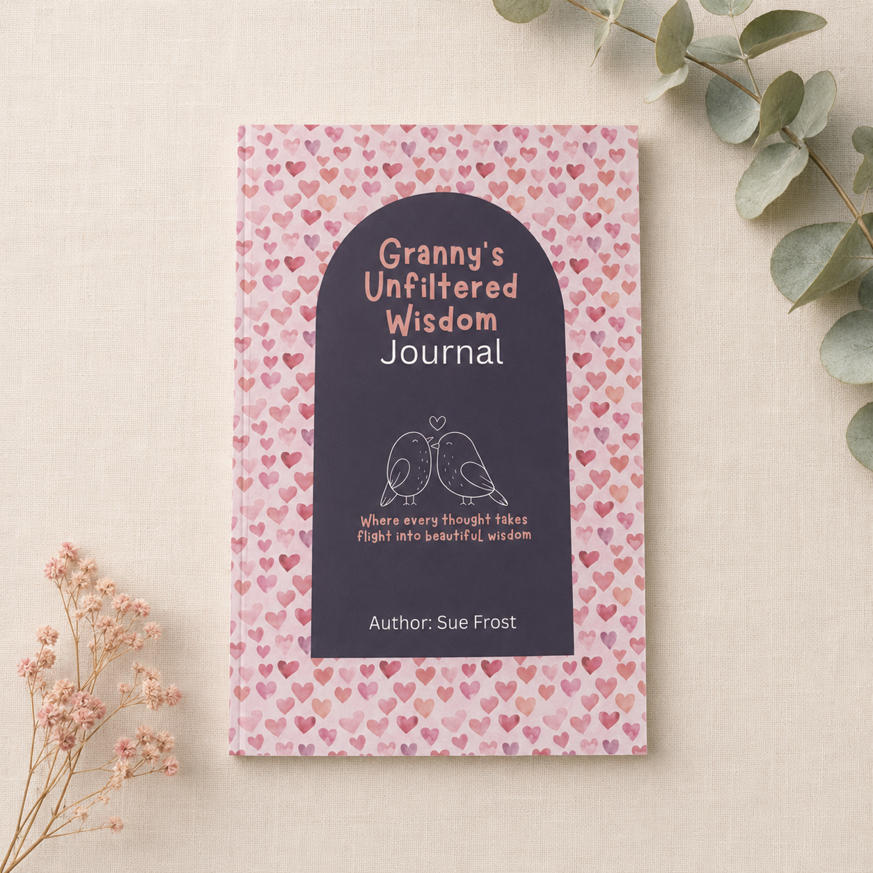 A journal titled 'Granny's Unfiltered Wisdom Journal' by Sue Frost, featuring a pink cover patterned with small hearts and a dark navy central label with white text and bird illustrations, styled on a beige fabric background with dried flowers and eucalyptus leaves.
