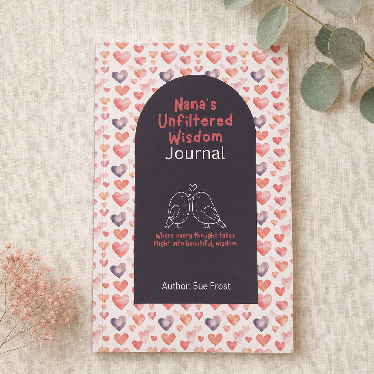 A hardcover journal titled 'Nana's Unfiltered Wisdom Journal' by Sue Frost, featuring a dark charcoal front cover with white and pink text and a bird illustration, set against a patterned pink and purple heart cover. The journal is styled on a beige linen surface with eucalyptus leaves and dried flowers.
