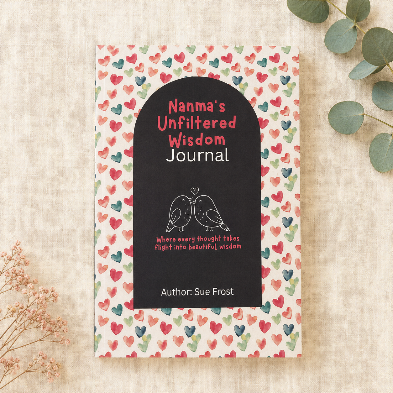 A hardcover journal titled 'Nanma's Unfiltered Wisdom Journal' by Sue Frost, featuring a black arched cover with white and pink text and a heart pattern border, styled on a beige fabric background with eucalyptus leaves.