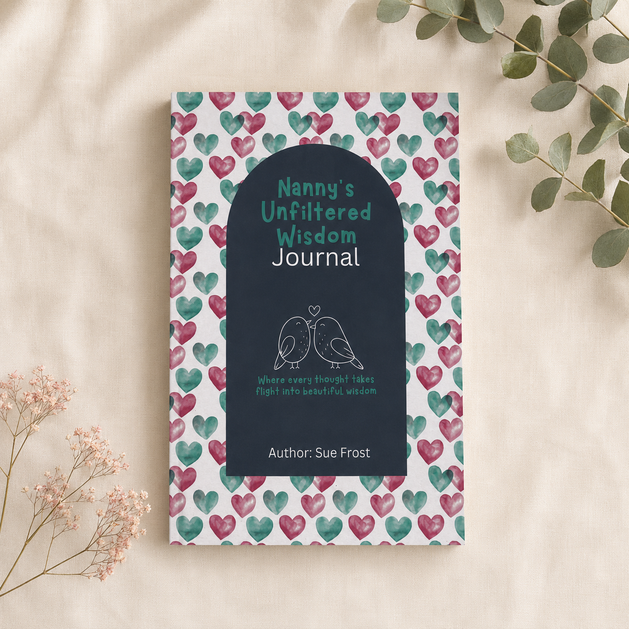 A journal titled 'Nanny's Unfiltered Wisdom Journal' by Sue Frost, featuring a dark navy blue cover with an arched central panel, set against a white background patterned with pink and teal hearts. The journal is styled in a flat-lay arrangement on a beige fabric background with eucalyptus leaves and small pink flowers.