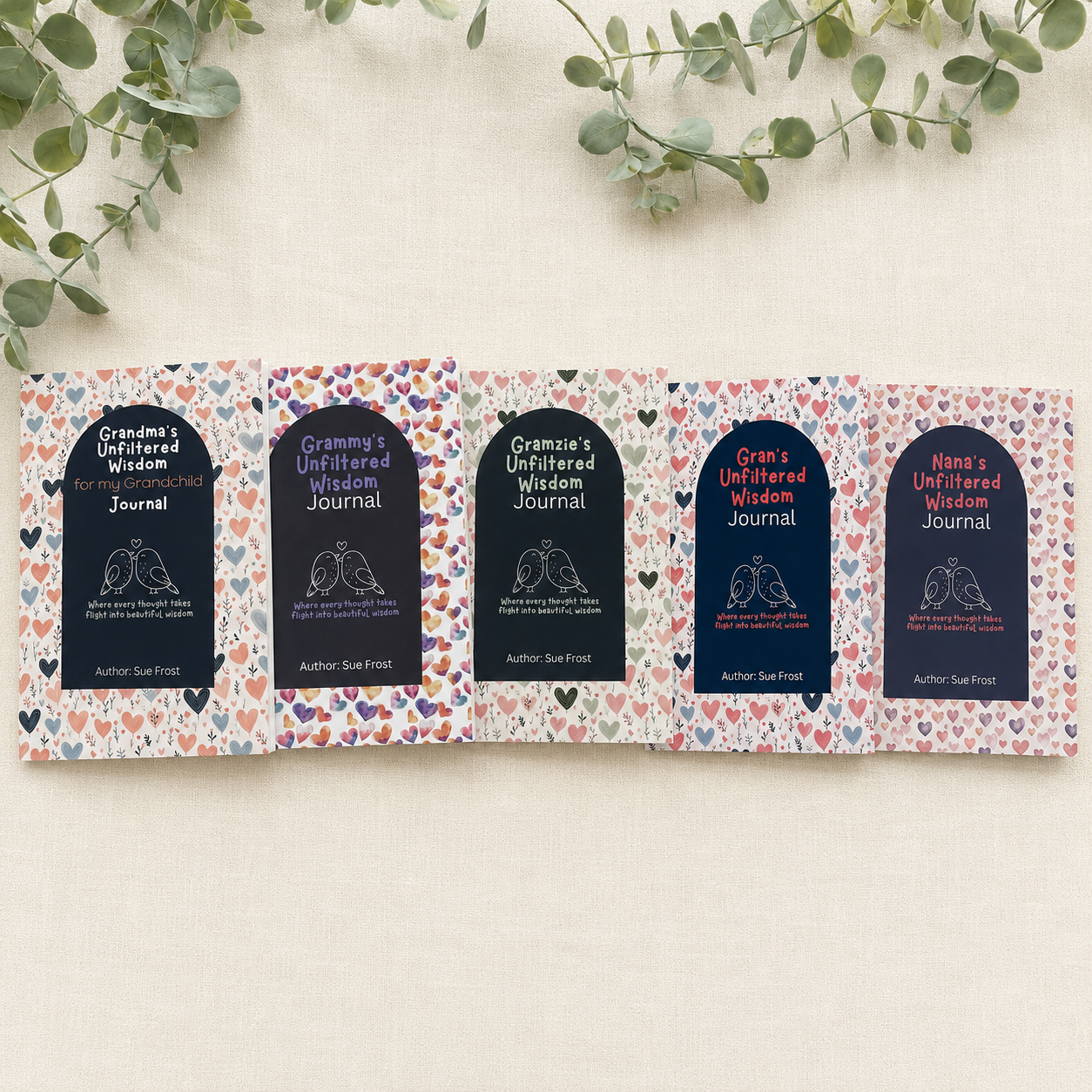Five folded pages of an unfiltered wisdom journal laid out on a light beige fabric background with green eucalyptus leaves at the top. Each page features a dark navy arch-shaped label with white text reading 'Grandma's Unfiltered Wisdom Journal' and 'Author: Sue Frost, surrounded by a colourful heart pattern. There is 4 other versions with Grammy, Gramzie, Gran and Nana.