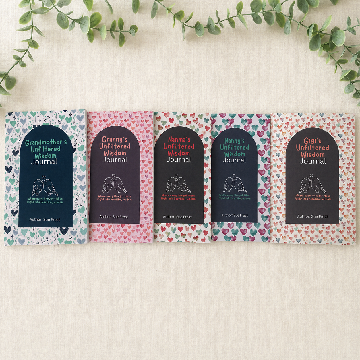 Five folded pages of an unfiltered wisdom journal laid out on a light beige fabric background with green eucalyptus leaves at the top. Each page features a dark navy arch-shaped label with white text reading 'Grandmother's Unfiltered Wisdom Journal' and 'Author: Sue Frost, surrounded by a colourful heart pattern. There is 4 other versions with Granny, Nanma, Nanny, Gigi.