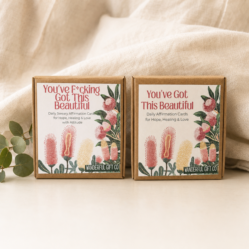 Two kraft paper gift boxes with floral affirmation cards, placed on a light surface with a draped beige fabric backdrop and eucalyptus leaves. The boxes are branded WANDERFUL GIFT CO and feature the phrases 'You've F*cking Got This Beautiful' and 'You've Got This Beautiful' with pink and yellow flowers.