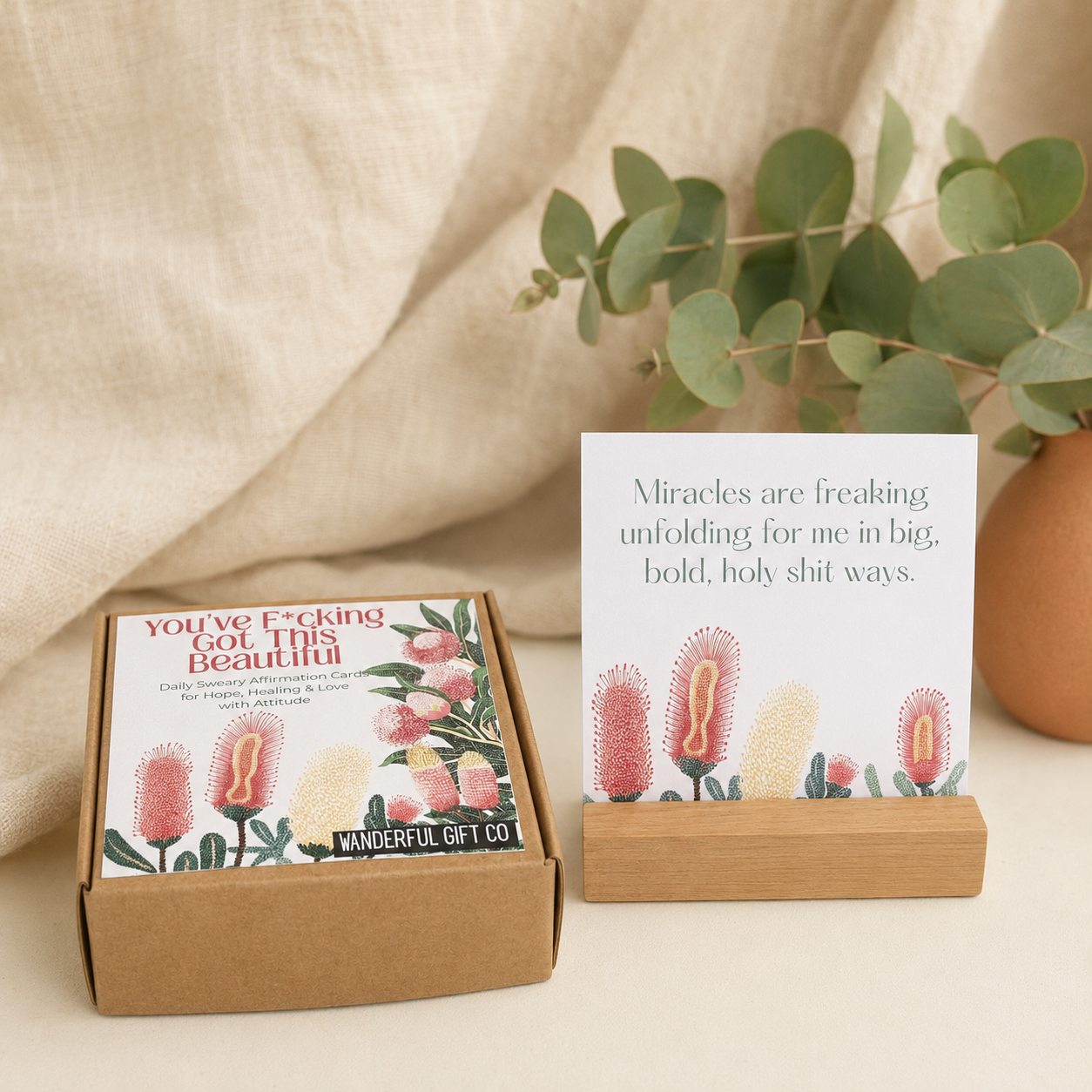 A kraft gift box containing a card and a small matching card stand, styled on a light surface with draped beige fabric and eucalyptus leaves in the background. The card reads, 'Miracles are freaking out for me in big, bold, holy shit ways.